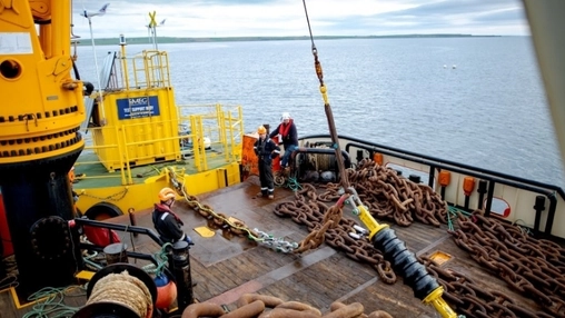 Workers in PPE managing rusted anchor chains beside yellow crane equipment on an IMEC offshore support vessel