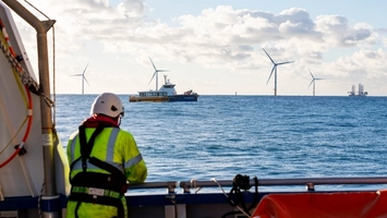 Technician in hi-vis jacket and helmet on a vessel deck overlooking an offshore wind farm