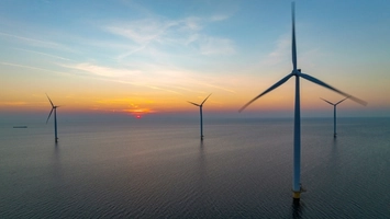 Four offshore wind turbines in calm open water at sunset with a glowing orange horizon and soft blue sky