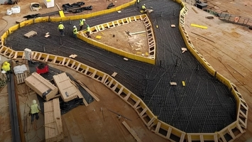 Large curved concrete foundation with yellow formwork, steel reinforcement mesh, and workers seen from above